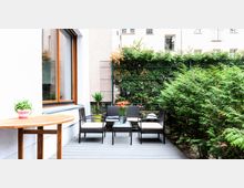 Kleine Terrasse mit Holzfußboden, Gartenmöbeln aus dunklem Geflecht mit hellen Polstern und einem runden Holztisch links. Im Hintergrund befinden sich grüne Hecken, ein Metallzaun und helle Hauswände mit Fenstern.