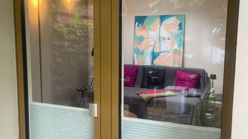 Blick durch eine zweiflügelige Glastür mit bronzefarbenem Rahmen und halbhohen, geriffelten Sichtschutzpaneelen. Dahinter ist ein Wohnraum mit Sofa, bunten Kissen, einem kleinen Tisch sowie einem großen abstrakten Wandbild und einem Dachfenster zu sehen.