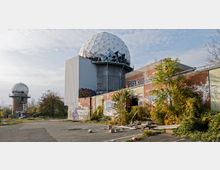 Verlassenes Gelände mit zwei Gebäuden; ein großer Turm mit einer geodätischen Kuppel obenauf und ein kleinerer, runder Turm links im Hintergrund. Die Bauten sind mit Graffiti bedeckt, umgeben von wuchernder Vegetation und verstreutem Schutt.