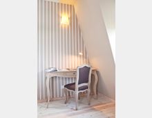 Holzschreibtisch mit geschwungenen Beinen und passendem Holzstuhl vor einer Wand mit vertikalen Streifen in Beige- und Weißtönen. Darüber befindet sich eine Wandlampe mit Schirm, die den Raum sanft beleuchtet.