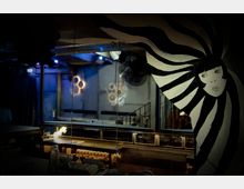 Innenansicht eines Bars oder Clubs mit industriellem Design. Im Vordergrund ist ein bemaltes Wandbild mit einem Gesicht und schwarz-weiß gestreiften Formen zu sehen, während im Hintergrund eine Theke, Treppen und Dekorationen mit Zahnrädern unter gedämpfter Beleuchtung erkennbar sind.