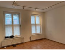 Ein leerer Raum mit hellen Wänden und Parkettboden, zwei große Fenster mit halbtransparenten weißen Gardinen, Heizkörper darunter und schwenkbaren Deckenstrahlern zur Beleuchtung. Außerhalb der Fenster sind Teile von Gebäudefassaden sichtbar.