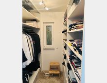 Ein kleiner begehbarer Kleiderschrank mit weißen Regalen auf beiden Seiten, die Kleidung und gefaltete Textilien enthalten. Am Ende des Raumes befindet sich ein Fenster, darunter ein Hocker auf einem beigefarbenen Teppich.