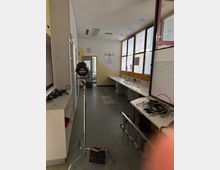 Ein schlicht eingerichteter Raum mit grauem Boden, großen Fenstern auf der rechten Seite und weißen Arbeitsflächen entlang der Wände. Im Vordergrund sind ein Stativ sowie technische Geräte und Kabel zu sehen, während eine Tür und eine Wandzeichnung (Vitruvianischer Mensch) den Hintergrund bilden.