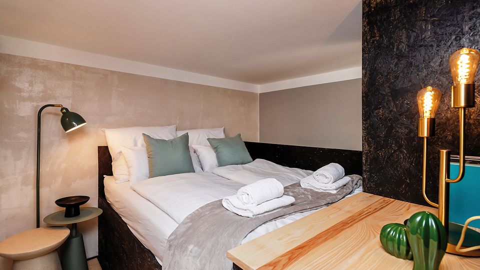 Modernes, kompaktes Schlafzimmer mit Doppelbett unter einer niedrigen Dachschräge, in neutralen Wandfarben und mit weißen Bettwaren, grauer Decke und zusammengerollten Handtüchern. Neben dem Bett stehen eine Stehlampe und kleine Beistelltische; im Vordergrund sind eine Holzablage und eine schwarze Trennwand mit warm leuchtenden Edison-Lampen zu sehen.