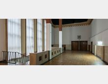 Großer Innenraum mit hohen, bodentiefen Fenstern, die von weißen Vorhängen bedeckt sind. Der Raum verfügt über einen Parkettboden, Holztüren an der Rückwand und eine helle Wandgestaltung mit dekorativer Bordüre unter der Decke.