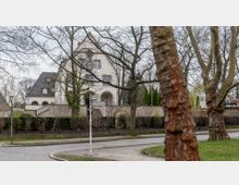 Straßenszene in einem Wohnviertel mit einer weißen Villa im Hintergrund, umgeben von einem Zaun und Bäumen. Im Vordergrund sind ein Wegweiser, große Baumstämme und eine gepflasterte Straße zu sehen.