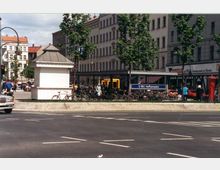 Straßenszene mit einer U-Bahn-Station („U-Bhf. Hermannplatz“) in Berlin, umgeben von mehrstöckigen Wohnhäusern im Hintergrund. Im Vordergrund eine kleine weiße Hütte, geparkte Fahrräder, Bäume und eine belebte Straße mit geparkten und fahrenden Autos.