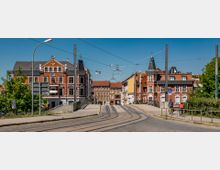 Straßenszene mit einer Brücke im Vordergrund und Gleisen für Straßenbahnen, umgeben von mehrstöckigen, historischen Gebäuden aus rotem Backstein mit weißen Details. Im Hintergrund sind Wohnhäuser und Fußgänger sowie Fahrradfahrer auf einem separaten Weg sichtbar, unter einem klaren blauen Himmel.