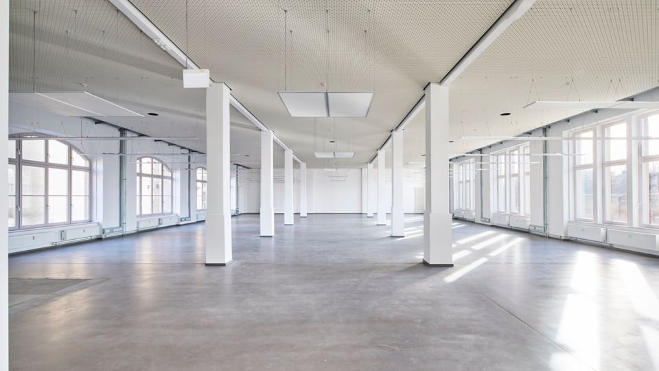 Weitläufiger, leerer Innenraum mit weißen Wänden und einer Reihe von rechteckigen Säulen. Der Raum hat große Fenster mit geschwungenen Oberlichtern, einen glatten Betonboden und eine perforierte Decke mit eingelassenen Leuchten.