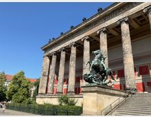 Außenansicht des Alten Museums in Berlin, mit seiner neoklassizistischen Fassade aus hohen Säulen und einem bronzenen Reiterdenkmal auf einem erhöhten Sockel im Vordergrund. Im Hintergrund sind rote und beige Wandpaneele sowie ein klarer blauer Himmel sichtbar.