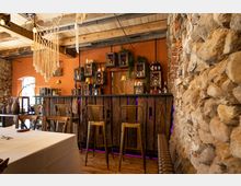 Rustikal gestalteter Barbereich mit hölzerner Theke, metallenen Barhockern und mit Pflanzendekoration geschmückten Wänden vor orangefarbenem Putz. Die Umgebung wird durch freiliegendes Mauerwerk und eine Decke mit Holzbalken ergänzt.