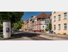 Straßenszene mit mehrstöckigen Wohn- und Geschäftshäusern in einer ruhigen Stadtumgebung, teils in klassischer und moderner Architektur. Links im Vordergrund steht eine Litfaßsäule, daneben sind Gehwege, Bäume und ein einspuriger Straßenabschnitt zu sehen.