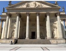 Neoklassizistische Kirchenfassade mit einem Dreiecksgiebel, verziert mit Reliefs und Statuen. Sechs korinthische Säulen tragen das Dach, und eine breite Treppe führt zum Haupteingang, der mit weiteren Skulpturen eingerahmt ist.