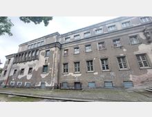 Ein großes, mehrstöckiges Gebäude mit verblasster, bröckelnder Fassade und zahlreichen Fenstern. Die Hauswand ist teilweise mit Graffiti bedeckt, und der Gehweg davor besteht aus Pflastersteinen.