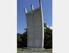 Ein großes, gewölbtes Betonmonument mit drei vertikalen Spitzen, umgeben von Bäumen und Blumenbeeten, vor einem klaren blauen Himmel.