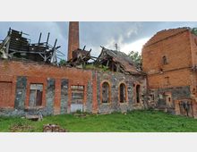 Ruine eines Backsteingebäudes mit teilweise eingestürztem Dach, gemischtem Mauerwerk aus Backstein und Feldsteinen sowie einem hohen Schornstein; im Vordergrund eine grasbewachsene Fläche.