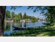 Flusslandschaft mit einem kleinen Boot im Vordergrund, umgeben von grüner Vegetation. Auf der gegenüberliegenden Uferseite stehen Häuser und Bäume, im Hintergrund ist eine Fußgängerbrücke zu sehen.