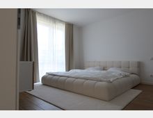 Modernes Schlafzimmer mit einem niedrigen, gepolsterten Bett in Beige, auf einem großen Teppich. Im Hintergrund befinden sich große Fenster mit lichtdurchlässigen weißen Vorhängen und beigen Verdunkelungsvorhängen, die viel Tageslicht hereinlassen.