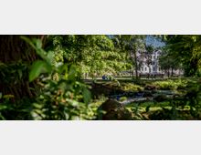 Ein grüner Park mit einem kleinen Bach, umgeben von Bäumen und Sträuchern. Im Hintergrund sind ein weißes Wohngebäude und vereinzelte Parkbänke zu sehen.