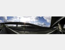 Panoramabild einer mehrspurigen Autobahn mit Verkehrsknotenpunkten und mehreren Brückenüberführungen. Der Himmel ist bewölkt mit Sonnenschein, und der Bereich ist von Betonbarrieren und modernen Straßenlaternen geprägt.