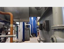 Industrieller Technikraum mit großen Rohrleitungen und einem blauen Druckbehälter im Hintergrund. Die Umgebung ist von Rohren, Leitungen, Ventilen und metallischen Geräten in einem engen, funktionalen Raum mit weißen Wänden geprägt.