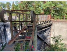 Alte, rostige Schleusenanlage mit einer metallenen Klappbrücke und massiven Betonwänden, umgeben von dichter Vegetation und einigen herbstlich gefärbten Bäumen.