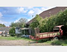 Ein ländlicher Hof mit einem roten landwirtschaftlichen Anhänger im Vordergrund, umgeben von überwucherten Büschen und Bäumen. Im Hintergrund stehen Gebäude mit roten Ziegeldächern und einigen gestapelten Materialien.