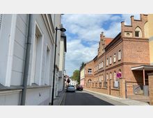 Schmale Stadtstraße mit parkenden Autos, flankiert von weißen Putzfassaden links und roten Backsteinfassaden im neugotischen Stil rechts. Im Hintergrund sind Bäume und weitere Gebäude unter einem bewölkten Himmel zu sehen.