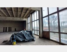 Ein leerstehender, industrieller Raum mit sichtbarer Betondecke und großen, raumhohen Metallfenstern, die den Blick auf Bäume und eine Außenfläche mit einem Gebäude freigeben. Im Vordergrund steht eine Plastikplane, bedeckt etwas auf dem Boden, neben einer Sprühflasche und einer Lampe.