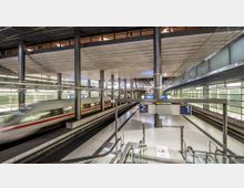 Moderne Bahnhofshalle mit zwei Gleisen, durchsichtigen Wänden und breiten Säulen. Ein Hochgeschwindigkeitszug passiert das linke Gleis, während oben eine Anzeige mit den Zahlen „2“ und „1“ hängt; Rolltreppen führen zu den Gleisen.