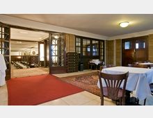 Ein Innenraum mit Restaurant- und Veranstaltungsbereich: Im Vordergrund stehen Holztische mit weißen Tischdecken, dahinter führt ein roter Teppich durch eine Glastür in einen weiteren Raum mit Stuhlreihen und Teppichboden.