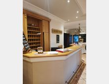 Hotelrezeption mit einem langen Tresen aus Holz, dahinter ein Regal mit Postfächern aus Holz. An der Wand befindet sich ein Schild mit der Aufschrift "RECEPTION", und die Decke ist mit Stuckleisten und eingelassenen Deckenleuchten verziert. Im Hintergrund hängt ein Kronleuchter, und rechts ist eine Glastür zu sehen.