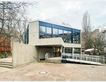 Zweistöckiges, modernistisches Gebäude aus Sichtbeton mit großen blau gerahmten Fensterfronten und einem überdachten Eingangsbereich. Davor liegt ein gepflasterter Vorplatz mit Treppen und einer barrierefreien Rampe mit Metallgeländer, umgeben von kahlen Bäumen und weiteren Gebäuden im Hintergrund.