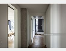 Flur eines modernen Bürogebäudes mit Holzboden und weißen Wänden, an dessen Ende ein Raum mit verglasten Fenstern und einer Staffelei sichtbar ist. Links ist ein Raum mit Konferenztisch und Stühlen zu sehen.