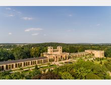 Historisches Gebäudeensemble mit symmetrischer Architektur, bestehend aus zwei Türmen und langen Wandelgängen mit Arkaden, eingebettet in eine parkähnliche Grünlandschaft; im Vordergrund gepflegte Gärten mit Brunnen, im Hintergrund eine waldreiche Umgebung unter klarem Himmel.