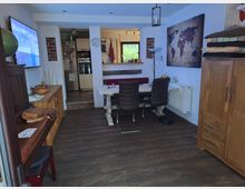 Wohnraum mit offener Küche und Essbereich, umfasst Holzmöbel, eine Weltkartenwanddekoration und einen Fernseher links an der Wand. Der Boden besteht aus dunklem Laminat, und der Zugang zur Küche erfolgt über eine Stufe mit Fliesen.