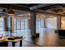 Ein modernes Loft mit großen Glasfenstern, sichtbaren Lüftungsrohren an der Decke und Betonpfeilern. Der Raum hat einen dunklen Fußboden, minimalistische Möbel und eine offene Gestaltung mit Blick nach draußen auf eine städtische Umgebung.