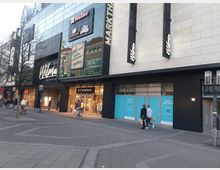 Einkaufszentrum mit moderner Glas- und Steinfassade, großer Schriftzug "Wilma Shoppen" und Schaufenstern von Geschäften, darunter "Eterna". Personen spazieren auf dem gepflasterten Gehweg, einige mit Einkaufstüten oder Fahrrädern.