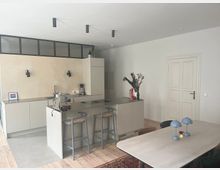 Moderne Küche mit hellen Fronten, einer Kücheninsel mit zwei Hockern und einer Arbeitsplatte mit Kaffeemaschine und Spüle. Im Raum befinden sich zudem ein Esstisch mit blauen Tischlampen, Holzfußboden und eine halbhohe Wand mit integrierten schwarzen Glasfenstern.