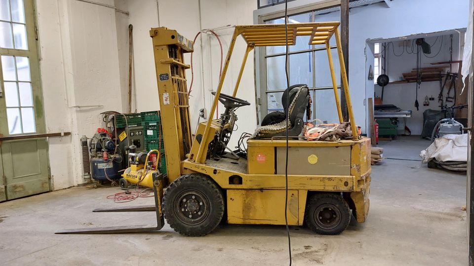 Ein gelber Gabelstapler steht in einer Werkstatt mit offenen Türen, Industriemaschinen und Werkzeugen im Hintergrund. Die Umgebung hat helle Wände, ein großes Fenster mit grünem Rahmen sowie teilweise sichtbare Lagergegenstände.