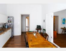 Helle Wohnküche mit einem großen Esstisch aus Holz in der Mitte, einer Küchenzeile mit weißen Schränken und einem silbernen Kühlschrank links. Im Hintergrund führen offene Türen zu einem Flur mit schwarz-weißem Fliesenboden und einem angrenzenden Raum mit Holzboden.