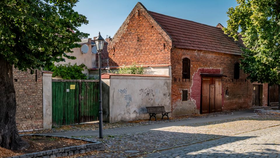 Kopfsteingepflasterte Straße in einem historischen Viertel mit einem niedrigen Backsteingebäude mit Satteldach, Holztür und kleinen Fenstern. Am Straßenrand stehen große Bäume, eine Laterne, eine Bank sowie eine grüne Holztorwand und angrenzende gemauerte Fassaden.