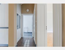 Schmaler Wohnungsflur mit beigefarbenen Wänden, weißen Türen und Holzfußboden mit dekorativem Fliesenstreifen in der Mitte. Am Ende führt eine Tür in einen hellen Raum mit grauen Bodenfliesen, seitlich öffnet sich ein weiterer Raum mit Parkettboden.