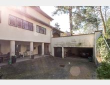 Ein gepflasterter Innenhof mit einem Wohngebäude im Hintergrund, das im oberen Stockwerk vergitterte Fenster und im Erdgeschoss eine überdachte Terrasse mit Säulen hat. Rechts befindet sich eine offene Doppelgarage mit Fahrrädern und einem geparkten Auto, umgeben von Bäumen und Efeu.