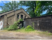 Backsteingebäude mit Ziergiebeln und seitlicher Holztreppe, daneben eine dunkelbraune Holzscheune mit großen Toren; umgeben von Pflanzen, Bäumen und etwas Unkraut auf dem Boden. Im Hintergrund blauer Himmel und grüne Baumkronen.