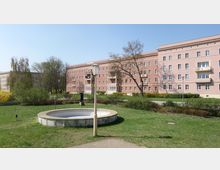 Wohnanlage mit rötlich verputzten vierstöckigen Gebäuden, Balkonen und symmetrischen Fenstern; im Vordergrund eine Grünfläche mit einem leeren Brunnenbecken, einer Laterne und Bäumen im Hintergrund.