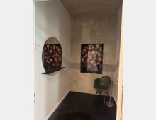 Kleiner Raum mit einer grauen Betonwand, einem dunklen Boden und einer weißen Seitenwand. An der Betonwand hängen Bilder von tulpenförmigen Blumen, und vor der Wand steht ein grüner Stuhl mit einer Stehlampe. Links ist ein ovaler Spiegel mit einem integrierten Holzregal befestigt.