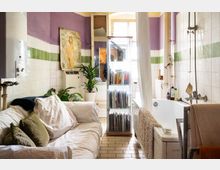 Ein Badezimmer mit weiß gekachelten Wänden, grüner und lila Bordüre, kombiniert mit einer Sofaecke. Eine Badewanne mit Duschvorhang befindet sich rechts, während links ein Sofa mit weißen Stoffen, grünen Kissen und Zimmerpflanzen steht. In der Mitte steht eine Glasvitrine mit Büchern.