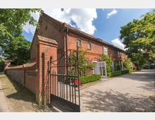 Backsteingebäude eines Landhofs mit Ziegeldach und weißen, bogenförmigen Fenstern. Davor befindet sich ein schmiedeeisernes Tor, ein gepflasterter Weg sowie gepflegte Hecken und Kletterpflanzen, umgeben von Bäumen.